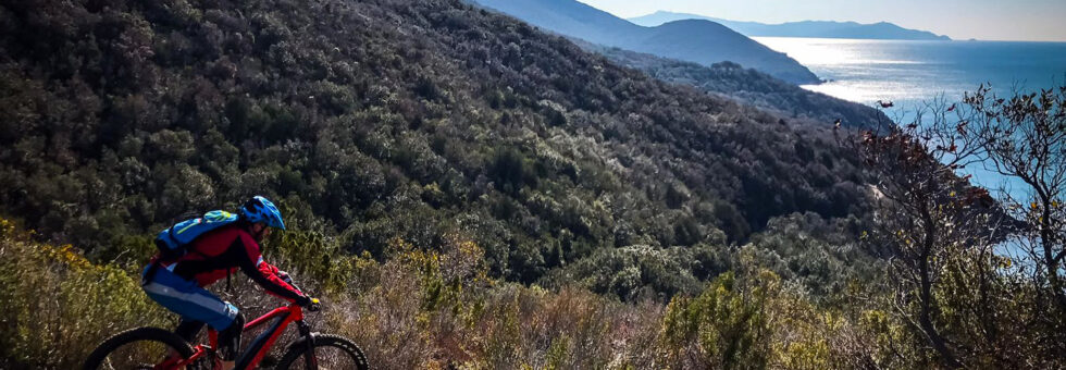 Un biker durante un'escursione in e-MTB enduro su un sentiero singletrack nella macchia mediterranea, con vista panoramica sul mare e sulle colline della Maremma toscana.