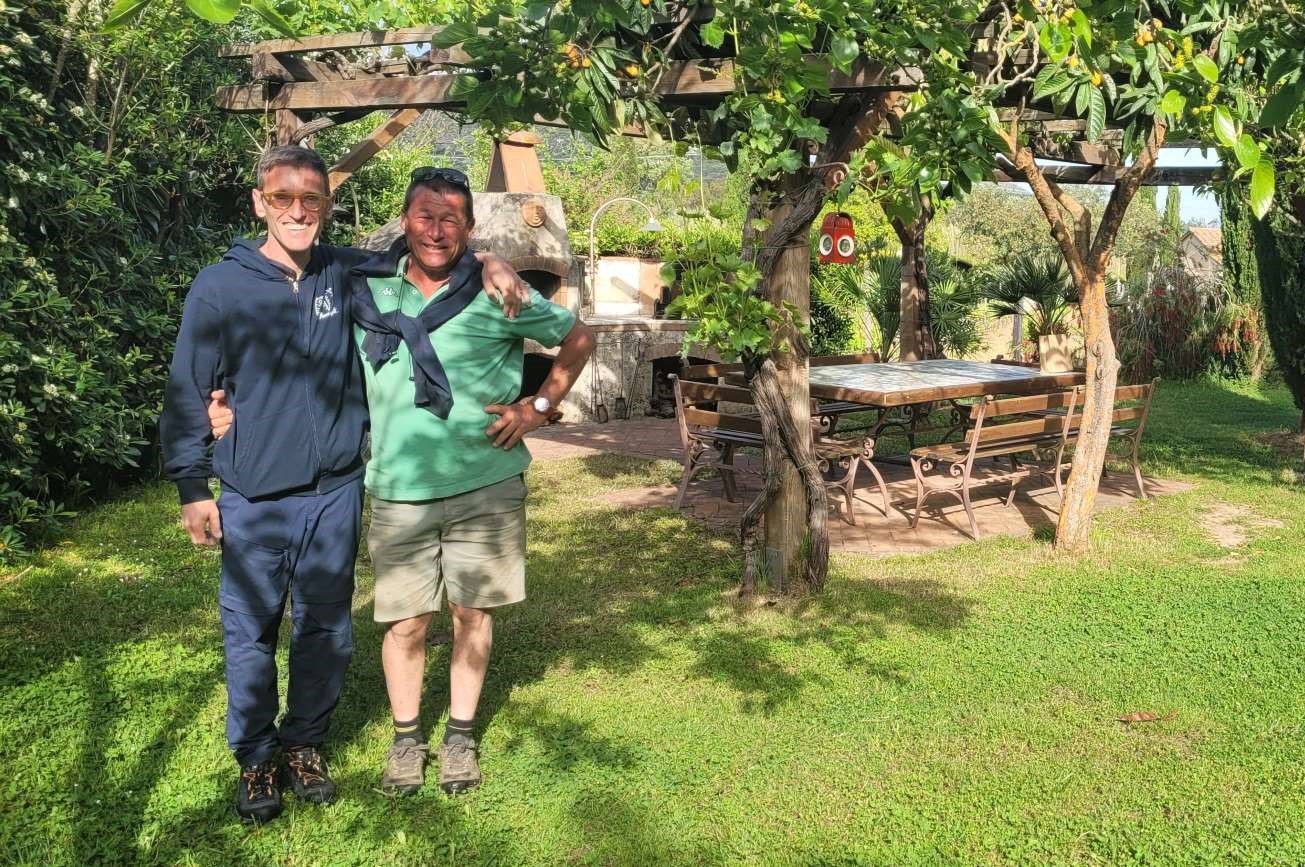Gianni, co-fondatore di Vacanze Sportive Toscana, abbraccia sorridente Eugenio Anselmi, il proprietario dell'Agriturismo Casa Marisa all'Isola d'Elba. I due si trovano nel curatissimo giardino verde della struttura, sotto una suggestiva pergola rustica che invita al relax dopo la fatica del trekking.