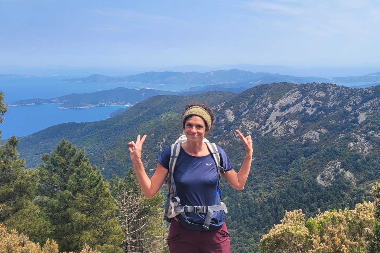 Patrizia in tenuta da trekking sorride facendo il segno della vittoria sul sentiero per il Monte Capanne all'Isola d'Elba, con il panorama verde e il mare dei golfi elbani sullo sfondo.