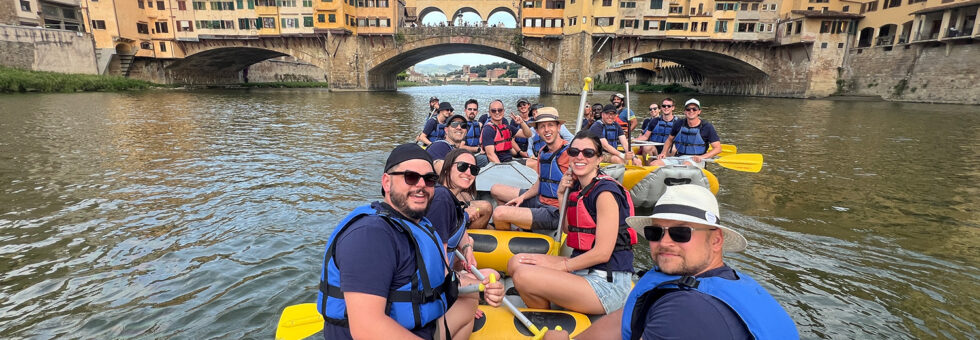 Un gruppo sorridente partecipa a un'escursione di soft rafting sull'Arno a Firenze, passando davanti a Ponte Vecchio con le guide di T-Rafting.