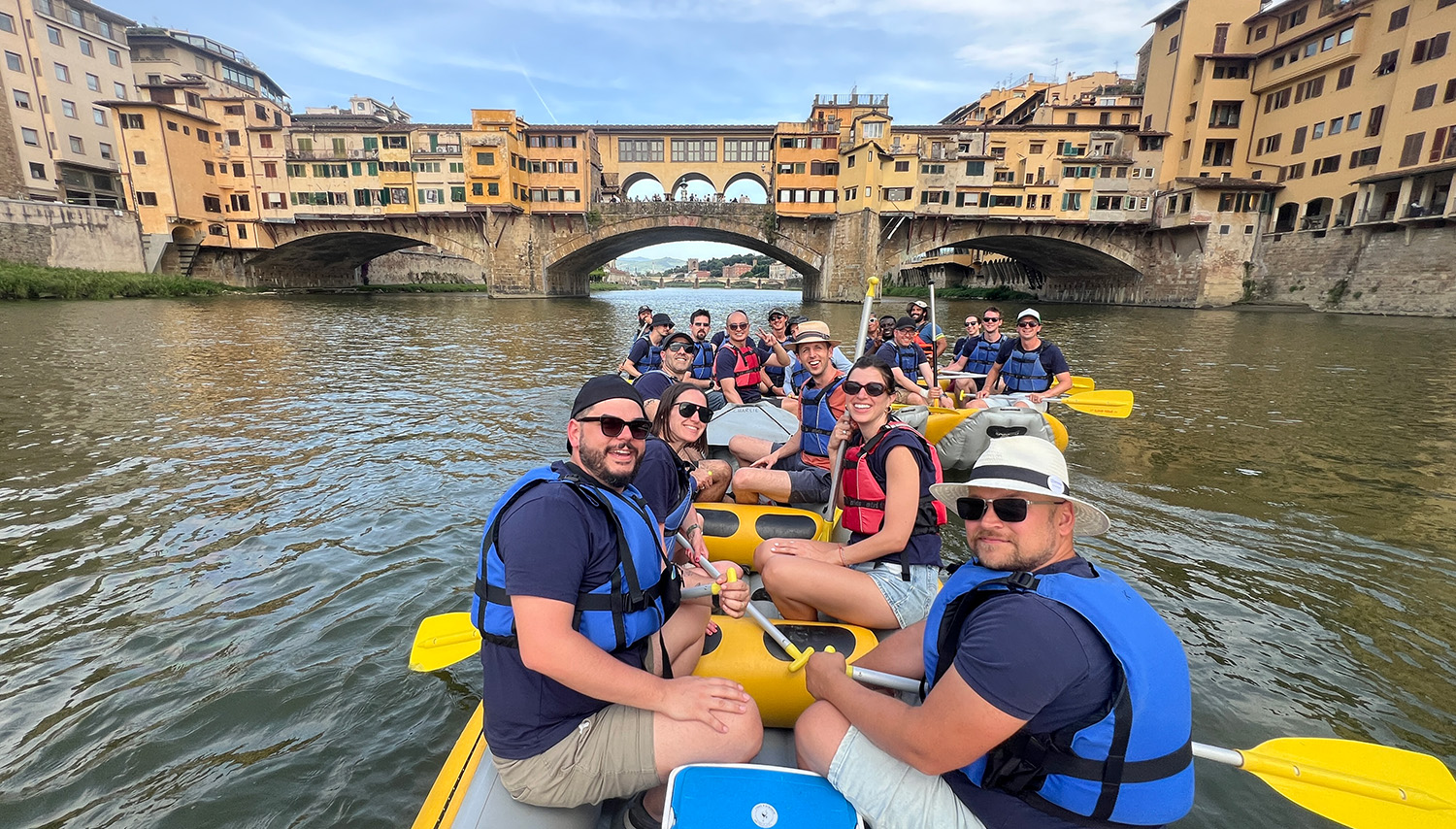 Un gruppo sorridente partecipa a un'escursione di soft rafting sull'Arno a Firenze, passando davanti a Ponte Vecchio con le guide di T-Rafting.